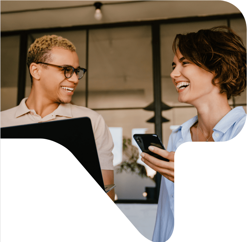 Two smiling people, one on a laptop and one holding a smartphone, look at each other.