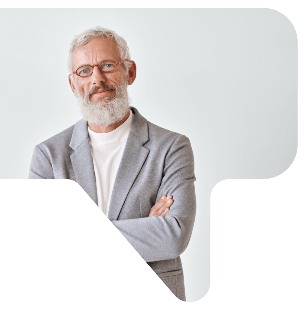 A grey-haired, bearded businessman in a white shirt and gray blazer wears red oval glasses with his arms crossed in front of a white, inside wall.