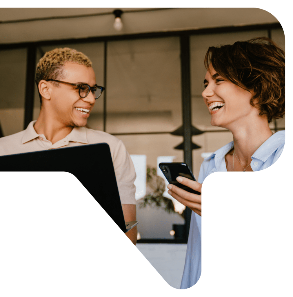 Two smiling people, one on a laptop and one holding a smartphone, look at each other.