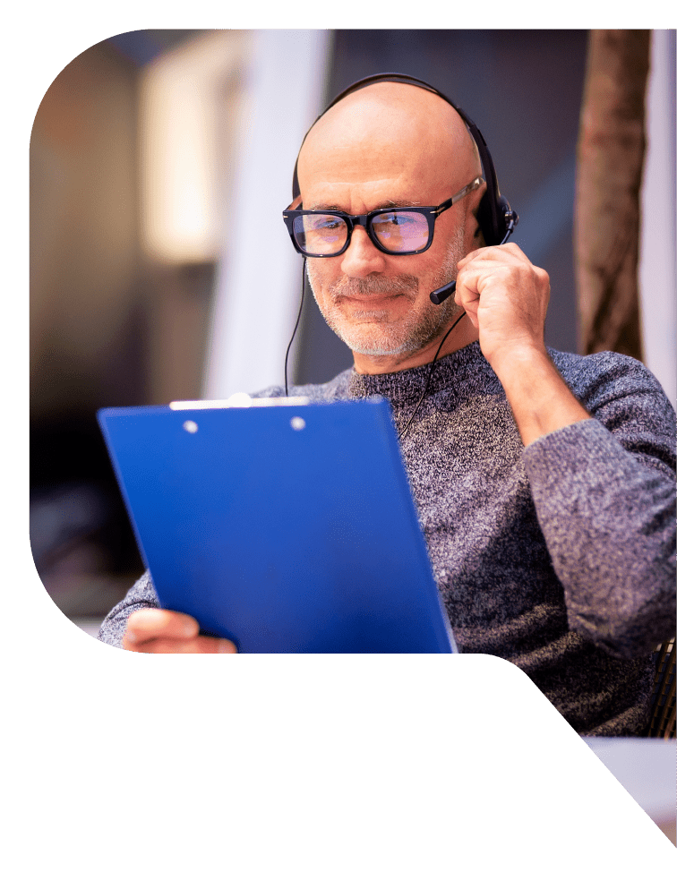 A bald man with glasses, wearing a headset looks at a blue clipboard that he's holding.