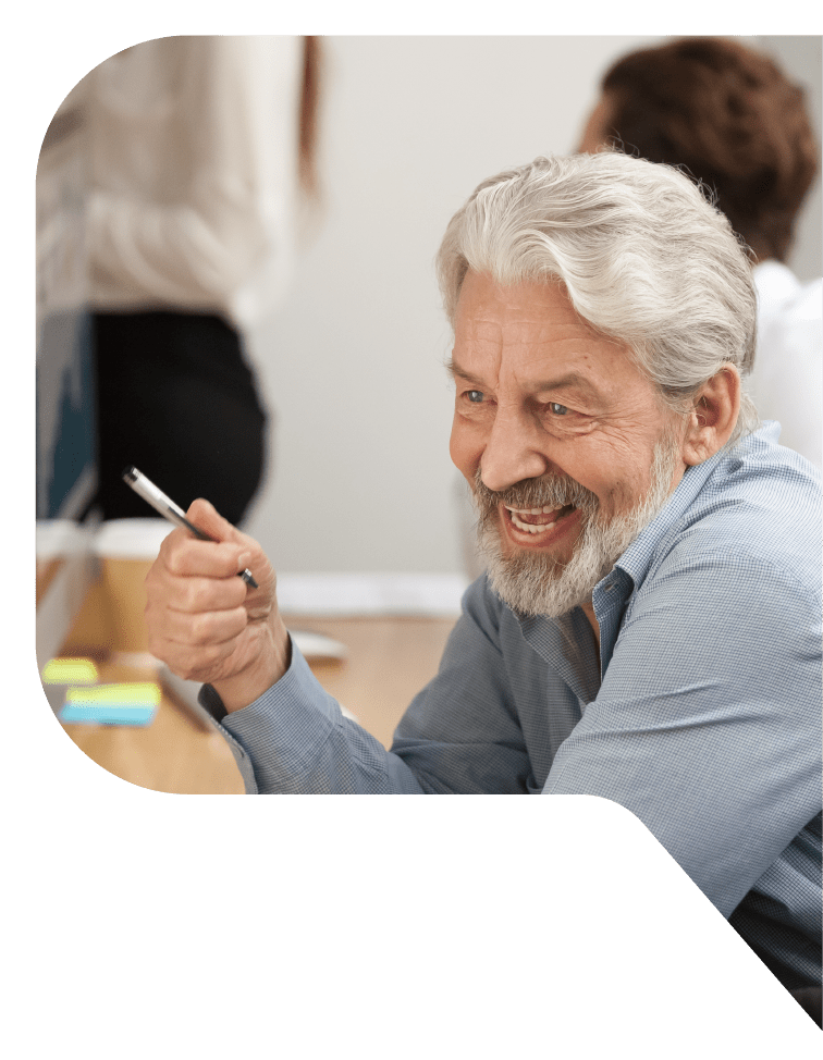 A smiling white-haired, bearded man holds a pen as he sits at a shared office desk with 2 people in the background.