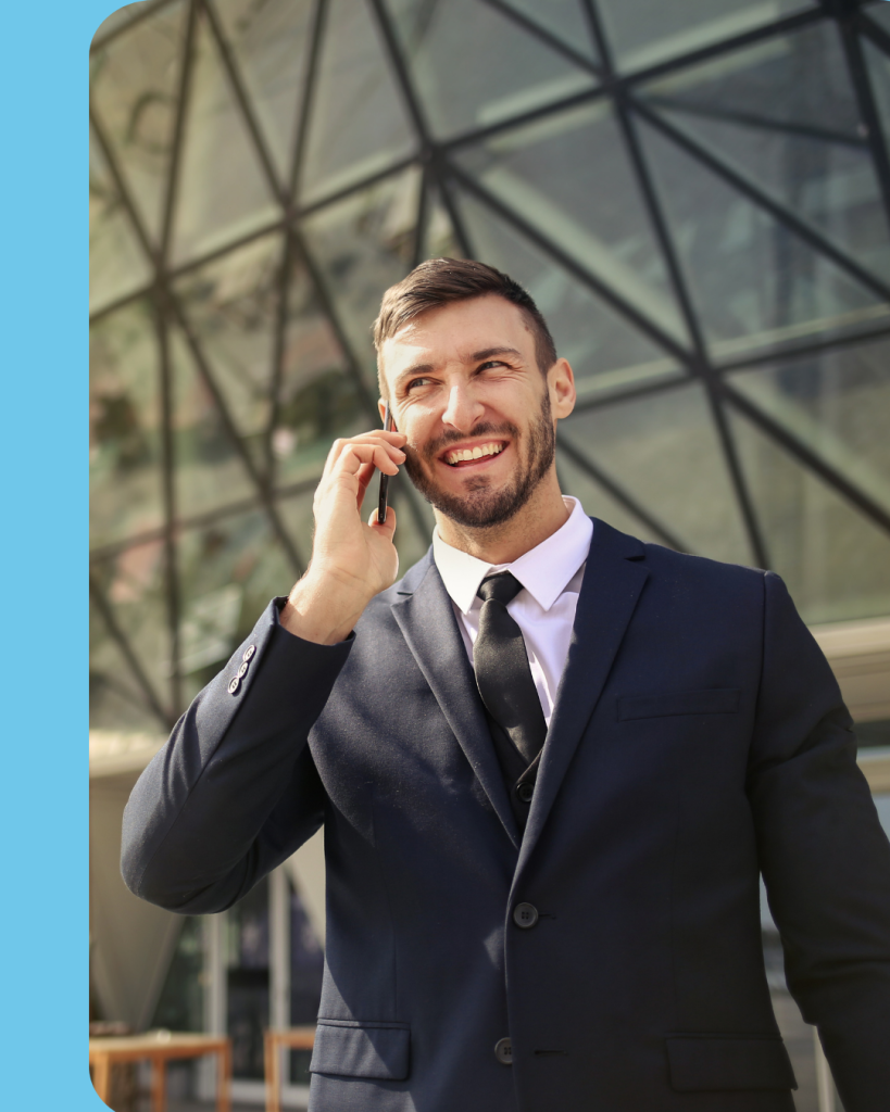 A smiling man looking up to the left outside of a modern glass building while holding a smart phone to his ear.