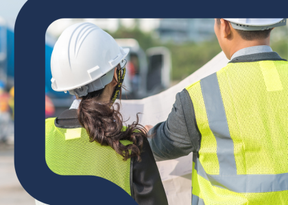 View from behind of a woman and a man in hardhats looking at a large blueprint outdoors.