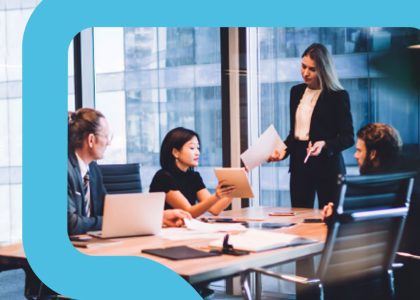 In a high, glass-windowed building, three people sit at a conference table showing information on a tablet to a standing woman.