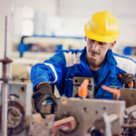 Factory worker in blue coveralls and a yellow hard hat operating a metal machine in a workshop with safety gear on