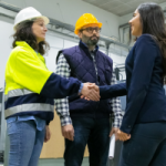 Two women and a man in safety gear shake hands in an industrial facility, signaling a meeting or agreement.