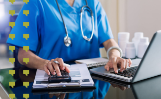 Healthcare professional in blue scrubs with a stethoscope uses a laptop and calculator at a desk, with a clipboard and papers nearby.